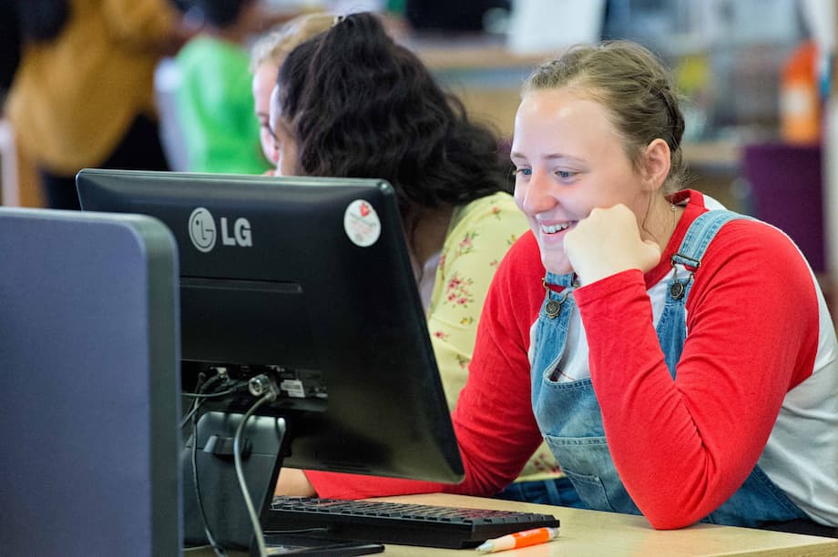 A woman sits at a computer in a library talking to friends.