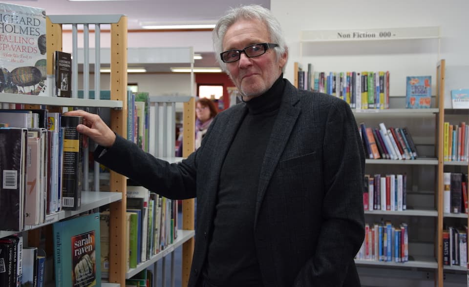 A man in jacket and glasses by a bookshelf.