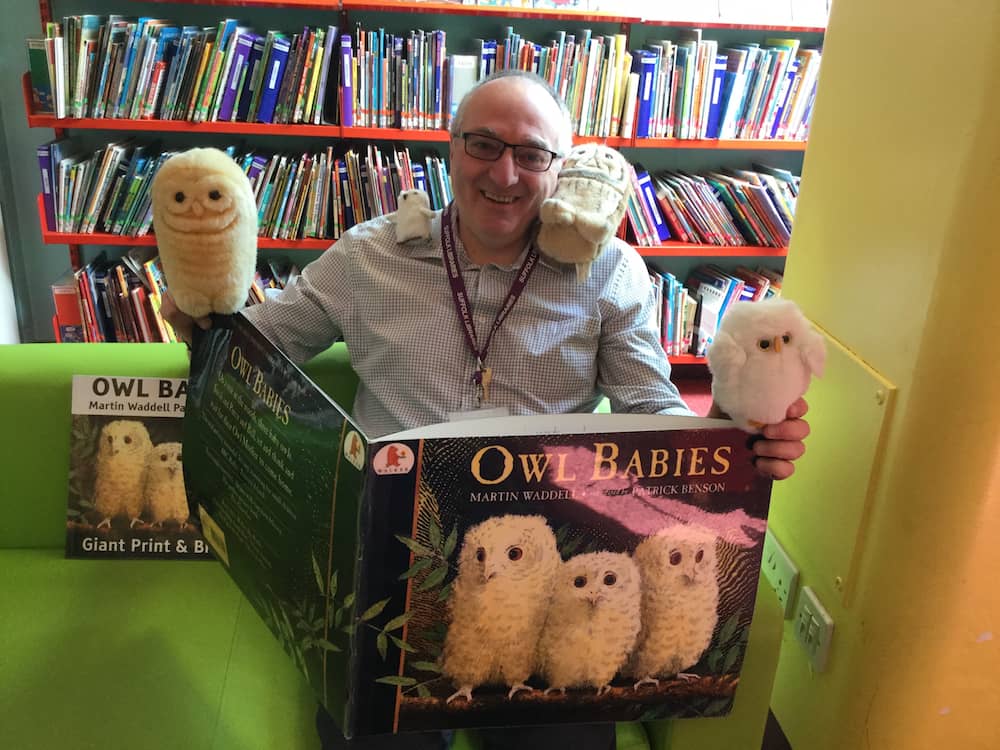 A man reading a huge hardback book with owl toys around him.