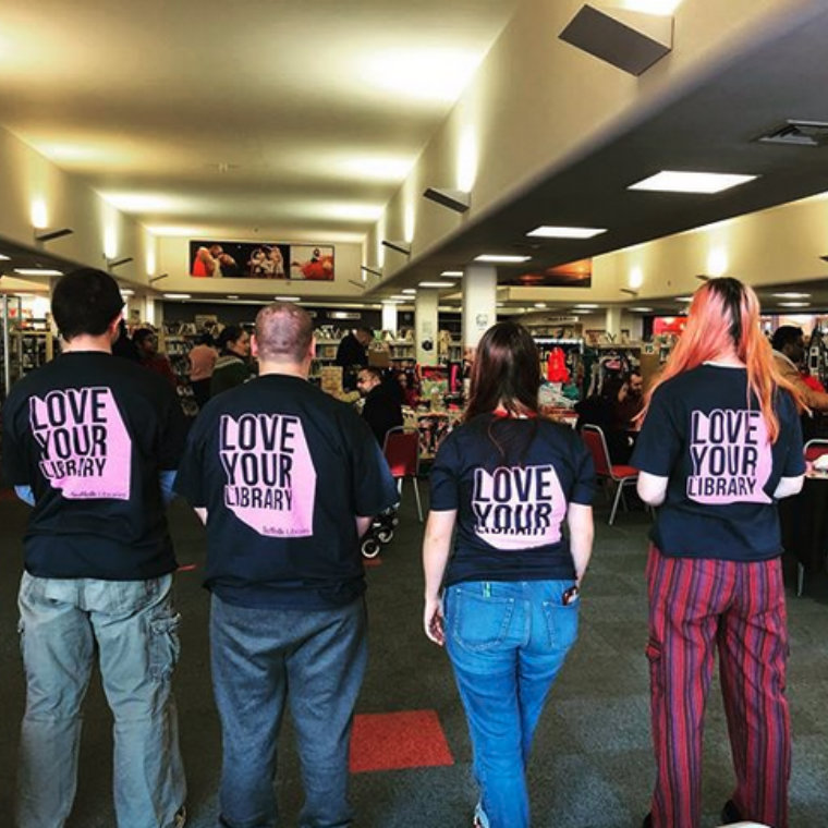 Four people in Ipswich County Library with their backs turned to the camera, wearing t-shirts reading 'Love Your Library'