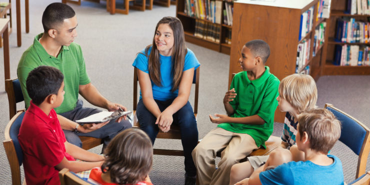 Teenagers in bright clothing sitting on chairs in a circle with a young man in a library