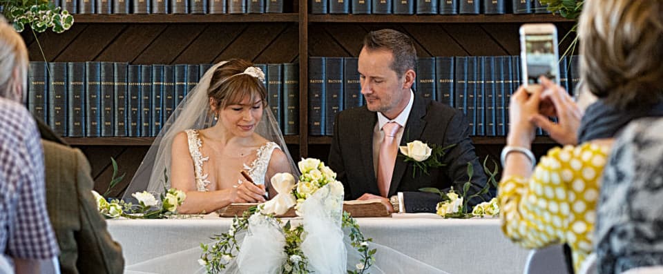 Two people signing a book at a desk in front of several witnesses.